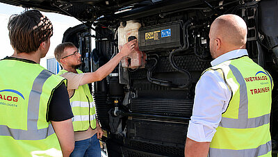 Wetralog-Trainer erklärt zwei Fahrschülern technische Komponenten unter der geöffneten Lkw-Front.