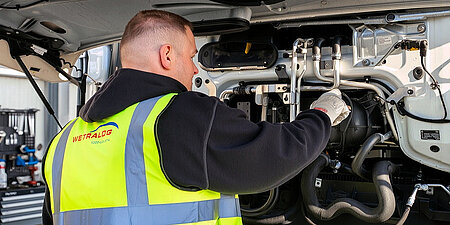 Wetralog-Mitarbeiter in Warnweste führt eine technische Kontrolle unter der geöffneten Lkw-Front durch.