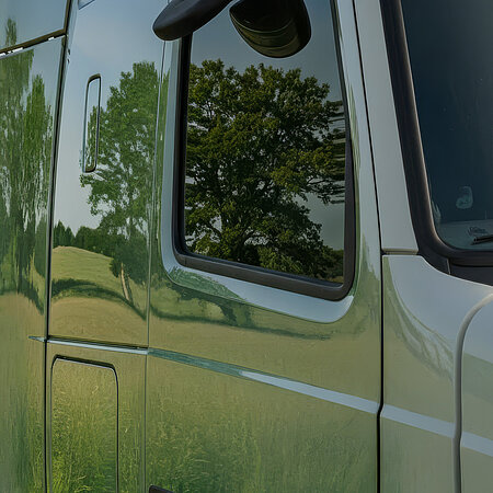 Nahaufnahme einer Lkw-Fahrerkabine, in deren Scheiben und Lack sich Bäume und eine grüne Landschaft spiegeln.