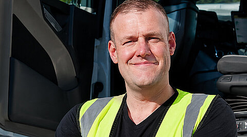 Lächelnder Lkw-Fahrer in Warnweste vor der geöffneten Fahrerkabine.