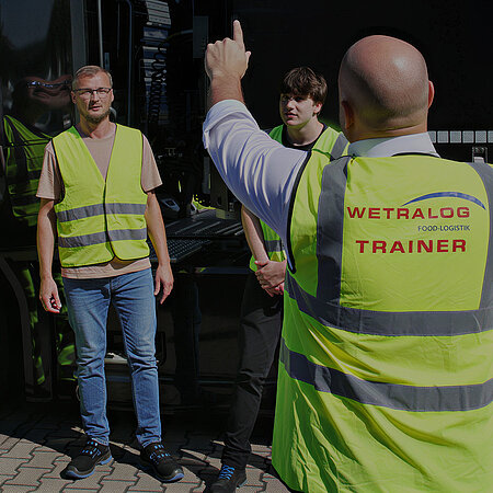 Wetralog-Trainer weist zwei Personen in Warnwesten an einem Lkw ein.