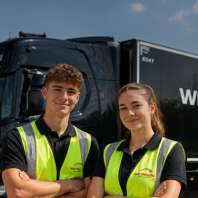 Zwei junge Wetralog-Mitarbeitende in Warnwesten stehen vor einem schwarzen Lkw.